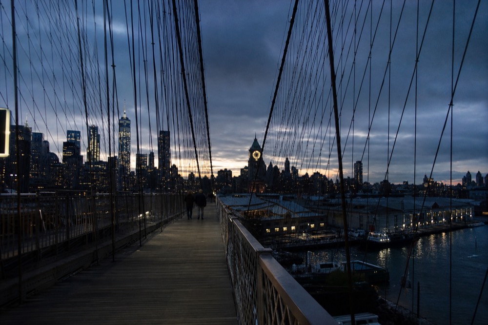 Suspension bridge cables converging above a wooden walkway, cold blue dusk behind, warehouse rooftops and a lit clock face on the far bank.