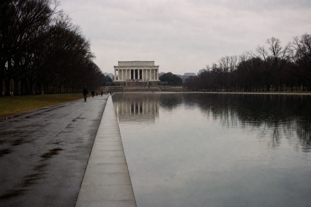 A wide neoclassical stone facade with columns at the end of a long reflecting pool, flat morning light, bare trees along both edges of the water.