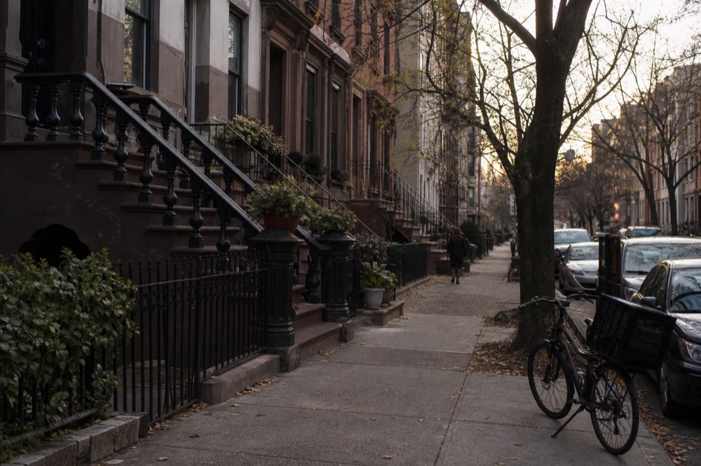 A quiet residential block of brownstone stoops in soft overcast light, iron railings, potted plants on the steps, one delivery bicycle leaning at the curb.