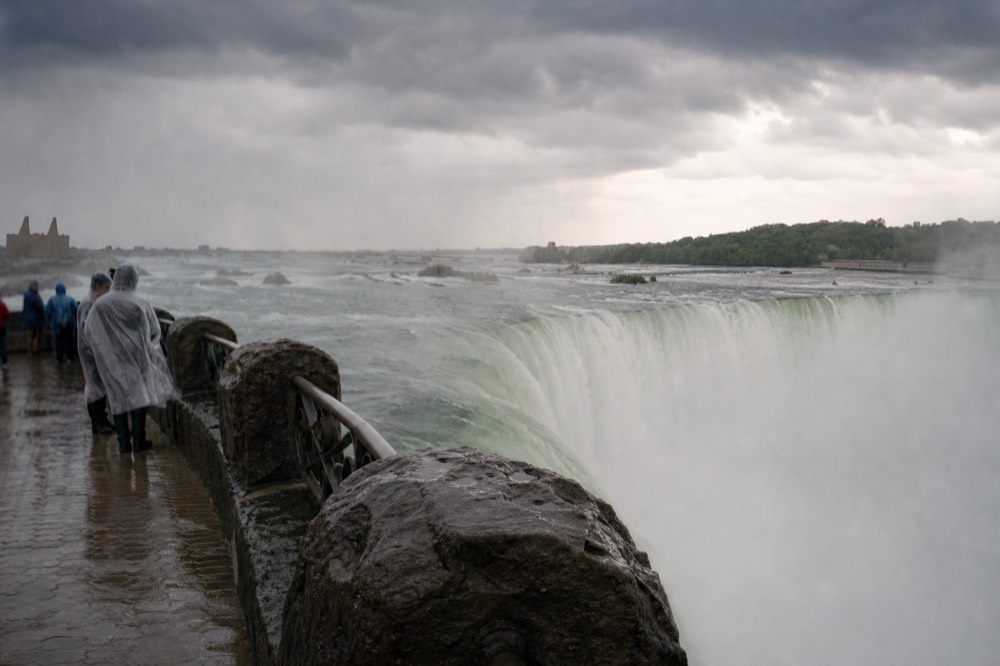 Green-white torrent pouring over a wide cliff edge seen from a mist-soaked railing, spray drifting sideways across wet stone decking under pale overcast sky