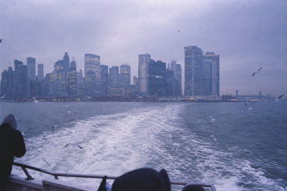 Harbor water churning white behind a sightseeing boat's stern at dusk, lower-city towers stacked in violet haze, gulls tracking the wake