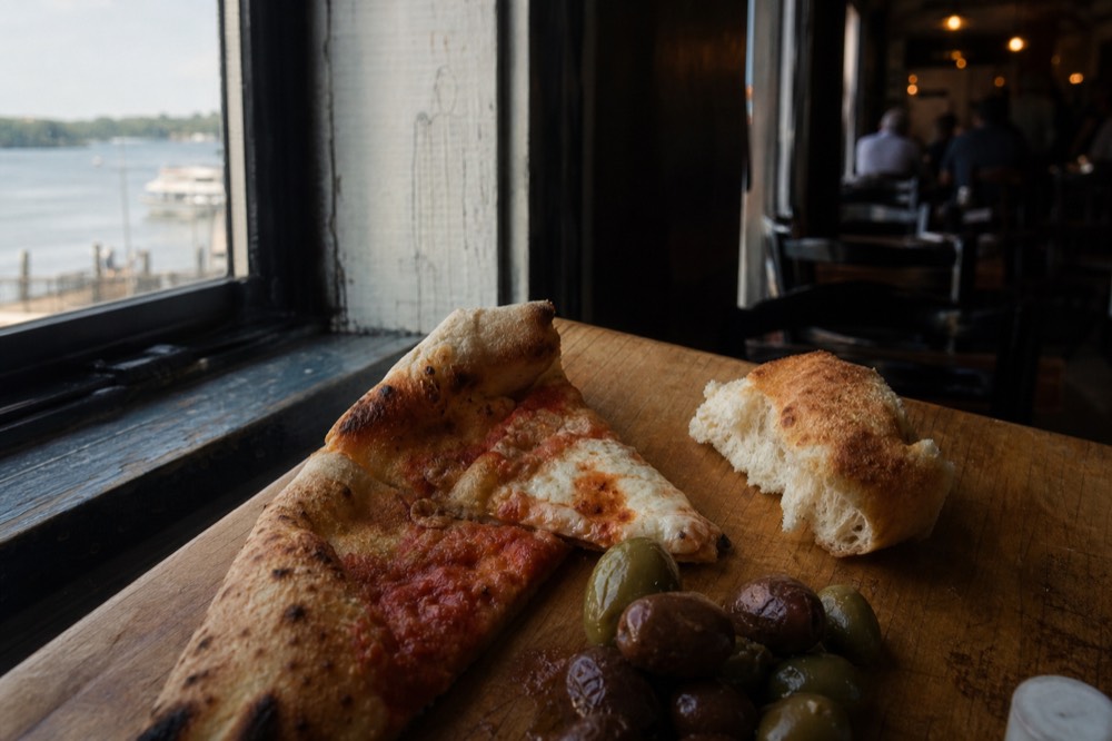 Close-up of a wooden board holding a folded pizza slice, torn bread and glossy marinated olives, soft window light from the left, no packaging or branding