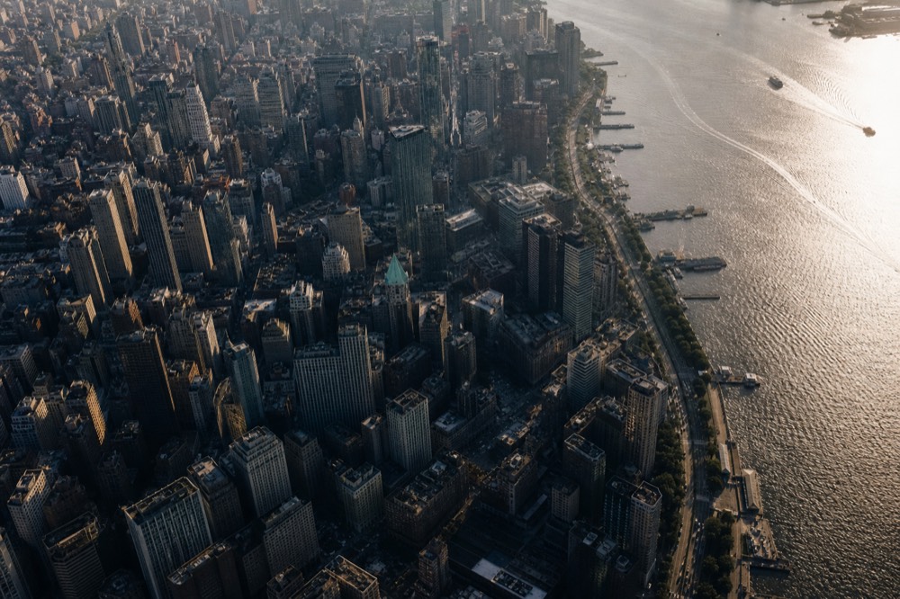 Dense grid of skyscrapers viewed steeply from the air in late afternoon, river glinting pewter along one edge, long shadows filling the avenues