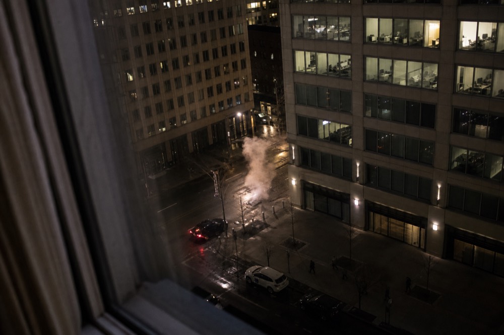Hotel room window looking down on a Midtown side street at night, wet pavement below, steam rising from a manhole, tail lights and lit windows opposite