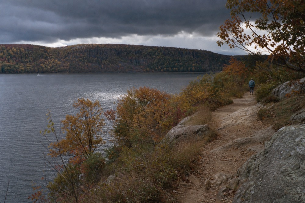 Forested ridge above a wide river gap in early autumn, patchy red and gold canopy, a narrow hiking trail of packed dirt and exposed grey rock