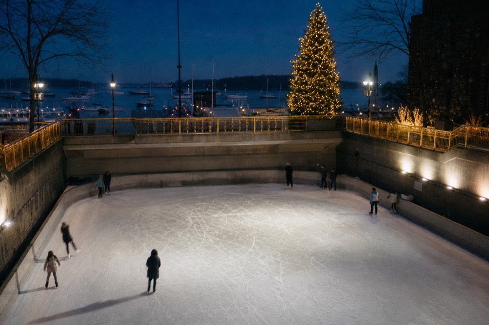 An open-air ice rink sunk below street level at night, skate marks scoring the ice, gilded terrace railings and a tall lit spruce rising behind the far wall