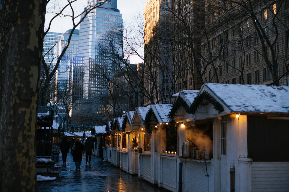 Rows of white wooden market stalls glowing at dusk under bare trees, steam rising from a food counter, snow dusting the tops of the canvas roofs