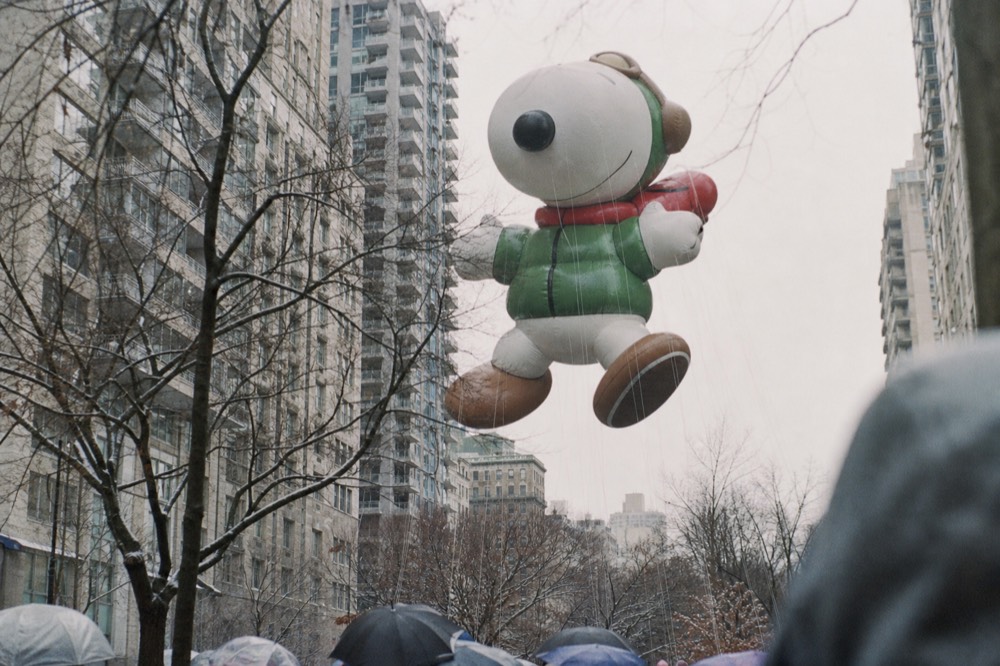A giant cartoon-shaped parade balloon floating between tall grey apartment buildings on a cold bright morning, handler ropes taut, bare branches in the foreground