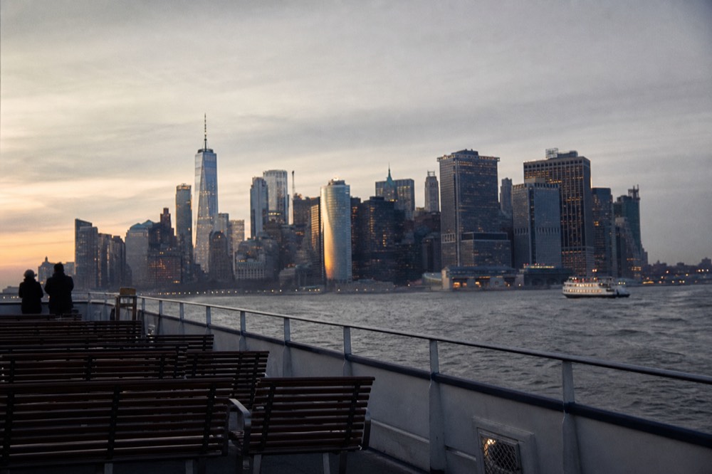 The open upper deck of a sightseeing boat on choppy grey harbour water in winter, wooden benches empty, low sun catching the towers of the skyline behind