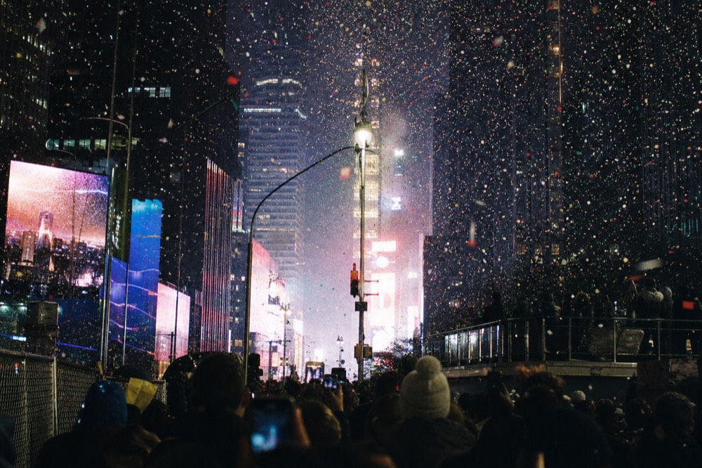 Confetti falling through the coloured glare of illuminated signs above a packed avenue at night, light streaking off metal barriers, no readable text on the screens