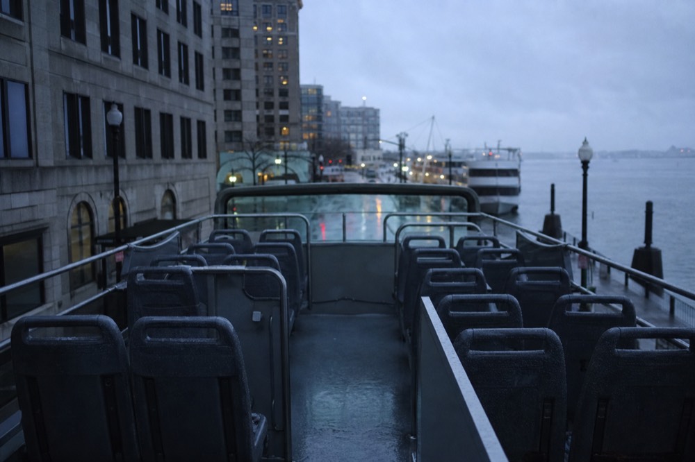 Empty upper deck of an open-top bus at street level, wet plastic seats and steel grab rails after a summer rain, grey overcast sky above