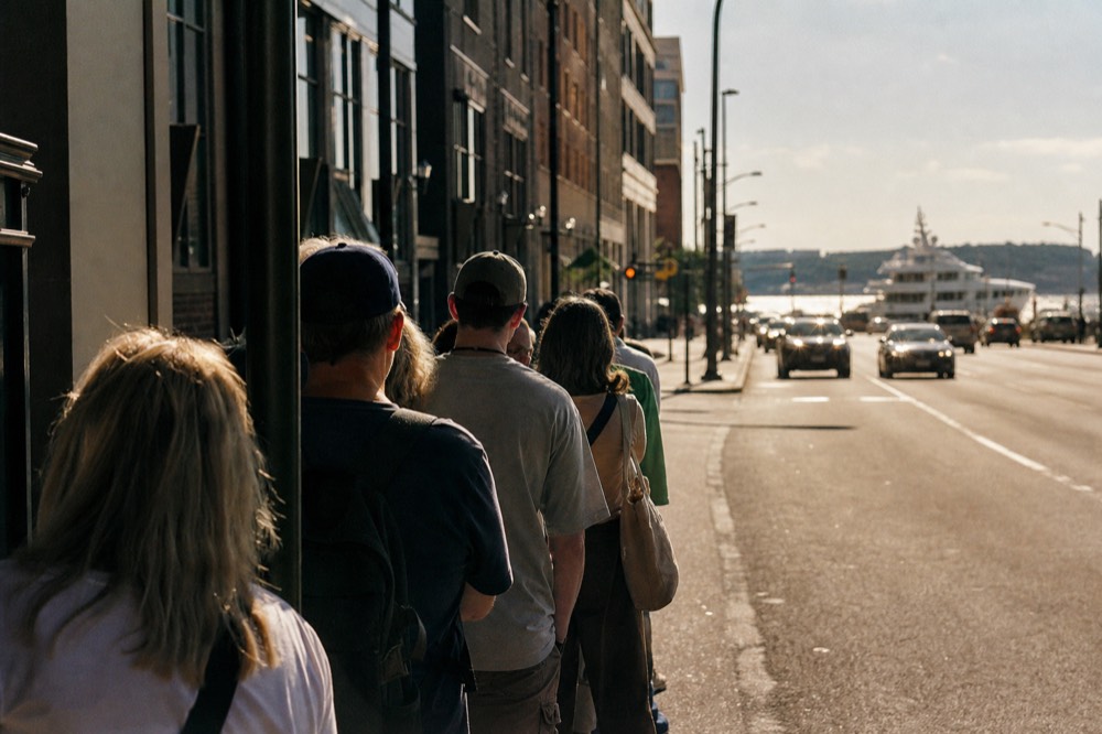 Line of travelers waiting at a curbside sightseeing stop beside a bare metal sign pole, shoulders hunched, hard midday glare bouncing off the sidewalk