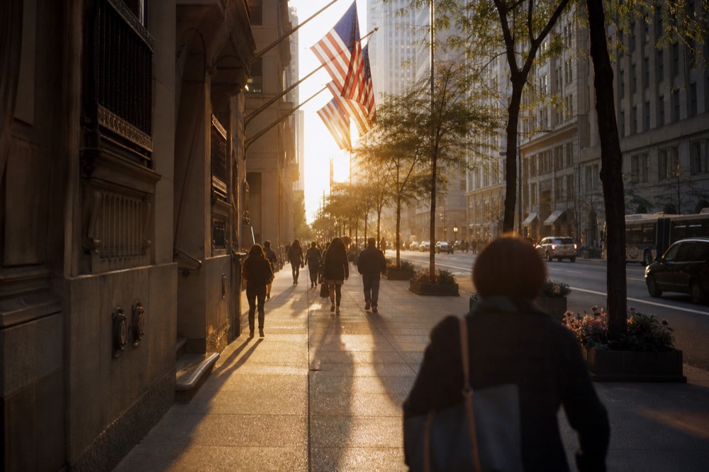 Wide Fifth Avenue sidewalk at golden hour, long pedestrian shadows stretched over paving stones, flags on limestone facades, no legible signage