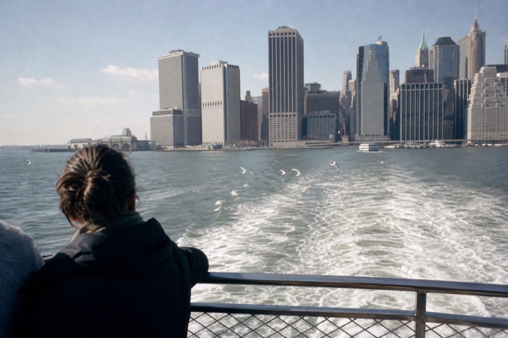 Harbor water churning white behind a sightseeing boat's stern rail, Lower Manhattan towers stacked in blue haze on the horizon, gulls trailing the wake