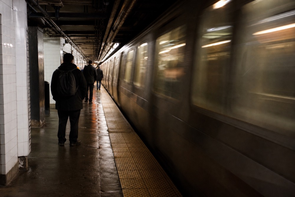 Downtown subway platform with white tiled walls and a train blurring past, warm tunnel light, a few commuters standing well behind the yellow edge line