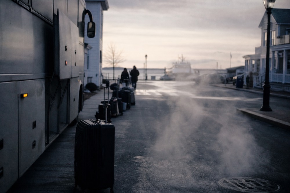 Motorcoach parked on a quiet hotel side street at dawn, suitcases lined along the curb, steam rising from a manhole grate under low grey light