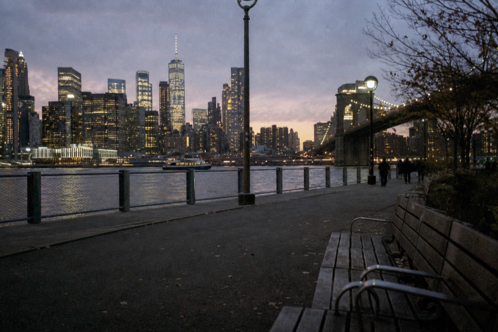 Brooklyn promenade path at dusk, bridge cables in silhouette, the lit skyline stretched across the East River, wooden benches empty in the foreground