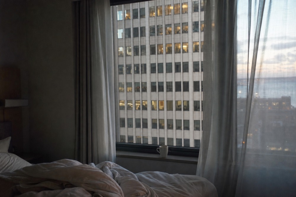 A hotel room at dawn with sheer curtains drawn back, an unmade bed, coffee cup on the sill and a wall of lit windows across the street beyond.