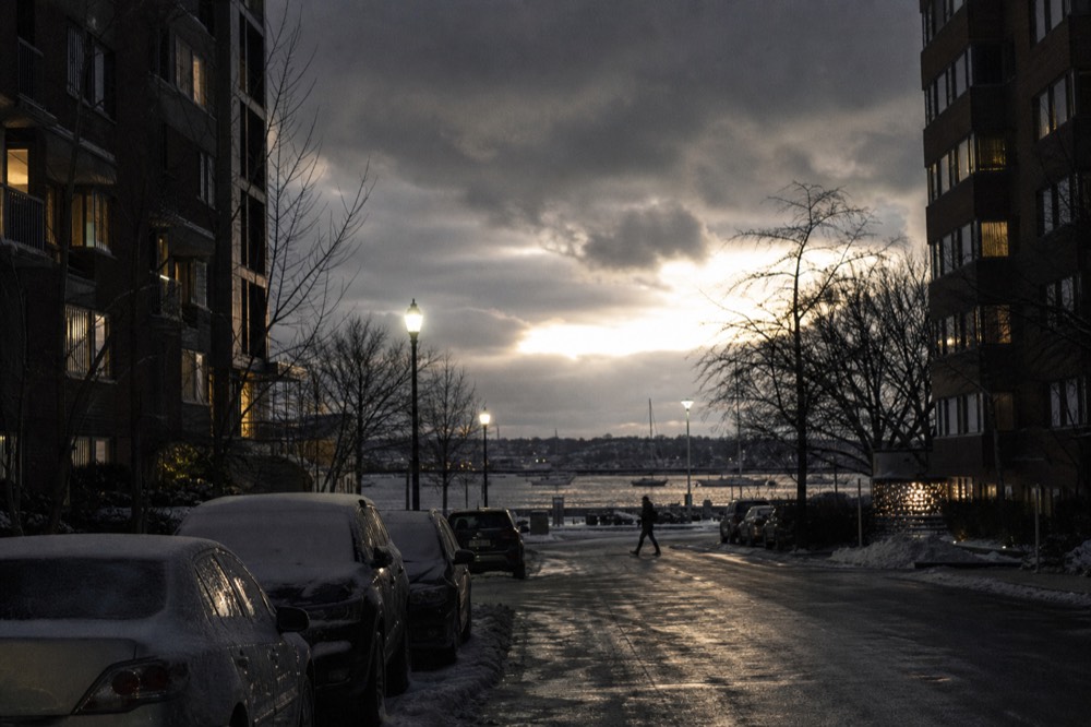 Snow settling on a row of parked cars beside a canyon of dark apartment buildings, warm yellow window light, a single figure crossing the empty street.