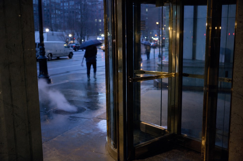 A revolving glass hotel door reflecting a rainy street, brass fittings catching lamplight, blurred umbrellas passing on the pavement outside.