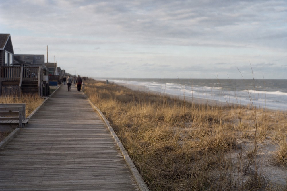 A weathered wooden boardwalk running beside pale dunes and marram grass, flat afternoon light, grey Atlantic surf breaking in the middle distance.