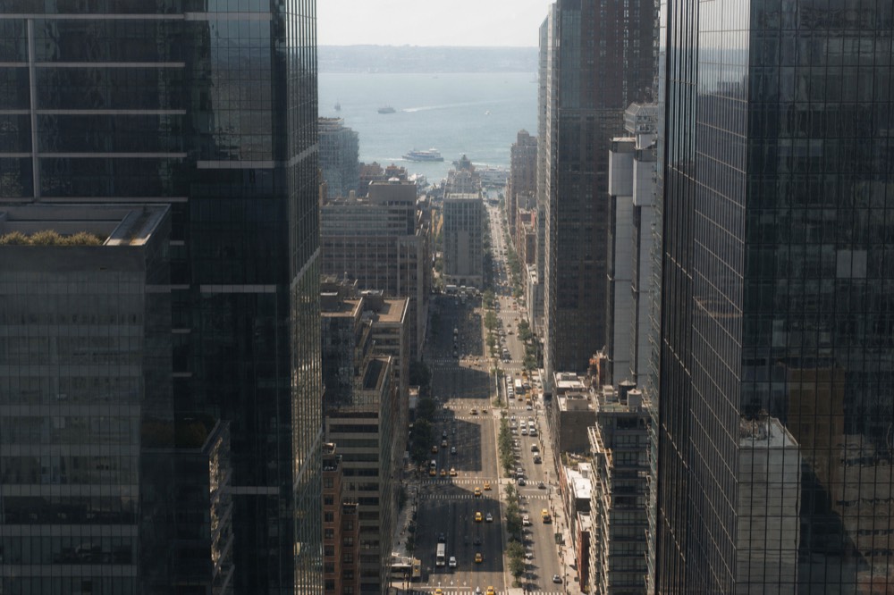 Looking straight down a canyon of glass towers from high above, tiny yellow vehicles threading the avenue, hard midday shadow cutting the street in half.