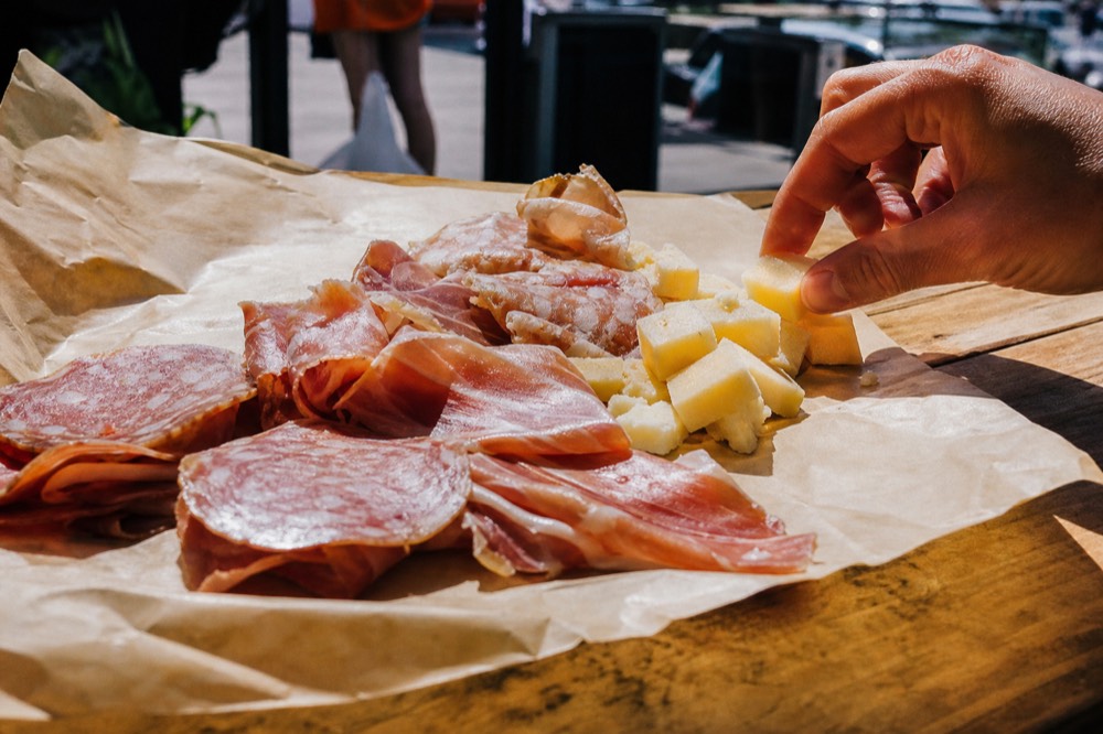 Close crop of sliced cured meats and cheese on butcher paper across a scarred wooden counter, warm overhead light, a hand reaching in from the frame edge.