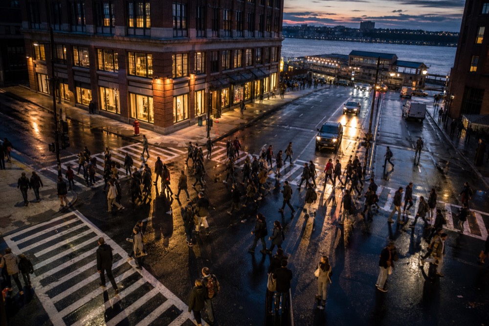 Overhead view of a crowded crosswalk at dusk, long shadows across white stripes, wet asphalt reflecting shop light, pedestrians mid-stride in every direction