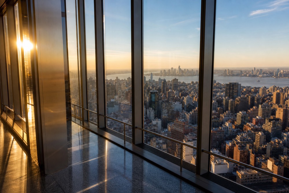 Glass-walled observation deck interior at golden hour, low sun flaring across polished floor, city grid stretching to a hazy river horizon, empty guardrail in foreground