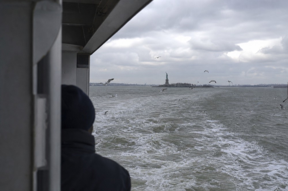 Harbor water churning green-grey behind a ferry stern, gulls tracking the wake, a small green statue silhouette on its island under flat cloud