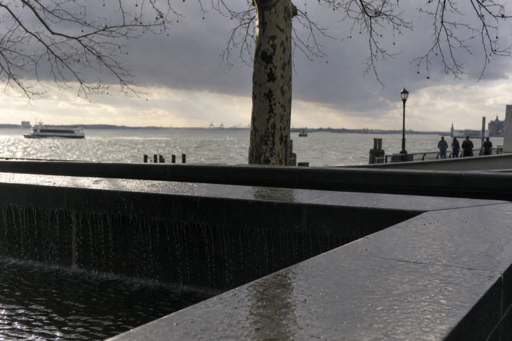 Water sheeting over dark polished granite into a square void, bronze parapet wet with spray, bare plane tree branches overhead against grey sky