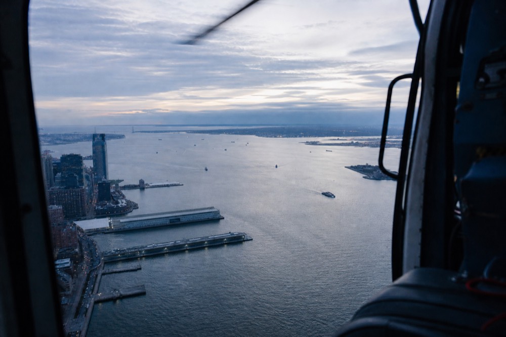 View through an open helicopter door frame over a wide river, rotor blur across the top edge, water glinting silver, piers and low warehouse roofs below