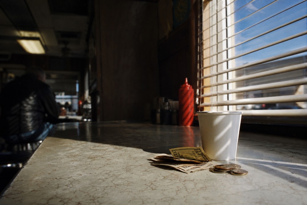 Folded bills and coins beside a paper coffee cup on a scratched diner counter, morning light through a slatted blind, ketchup bottle out of focus behind