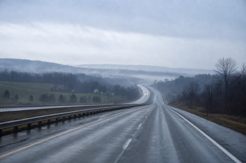 Empty interstate cutting through rolling upstate farmland at dawn, mist sitting in the hollows, guardrail and worn white lane markings receding toward low hills