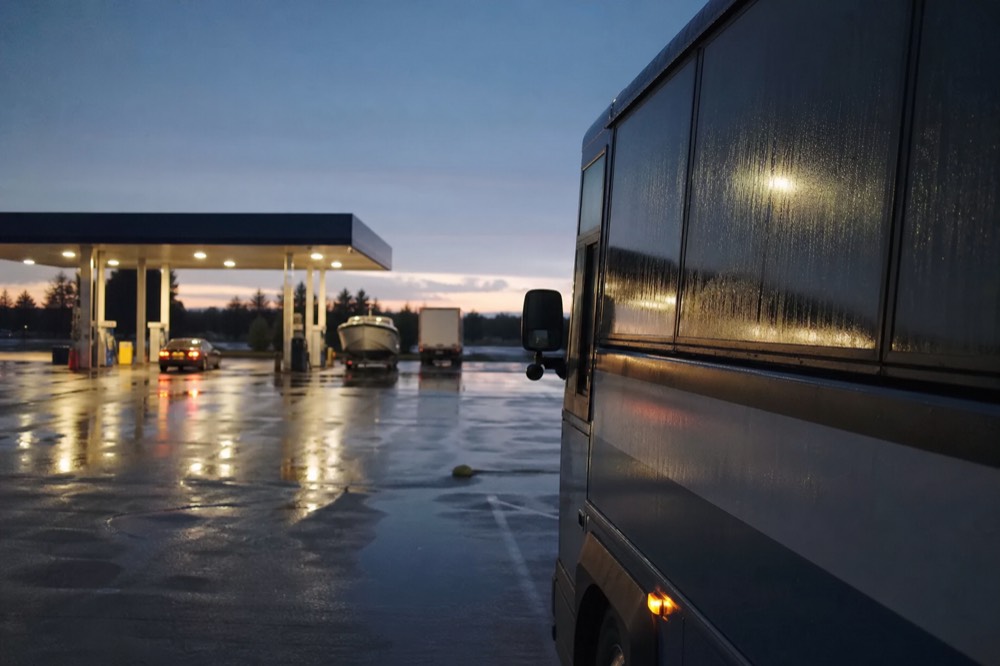 Rain-streaked coach windows at a highway service stop before sunrise, wet asphalt reflecting the fuel canopy lights, no signage or branding visible anywhere