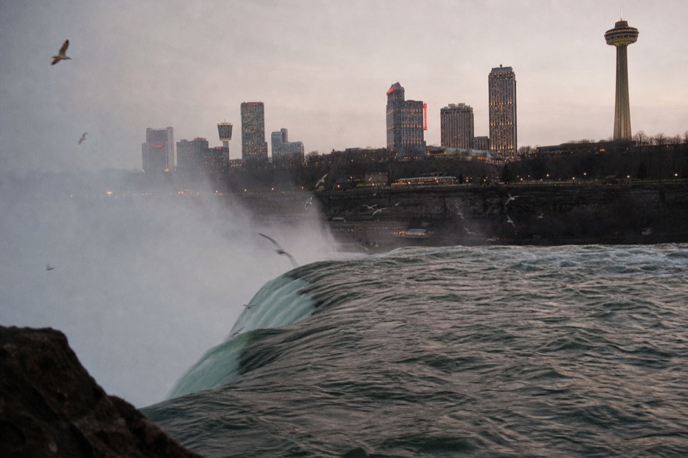 Green river water accelerating over a smooth stone lip into rising vapor, gulls circling above the drop, dark spray-soaked gorge walls on both sides