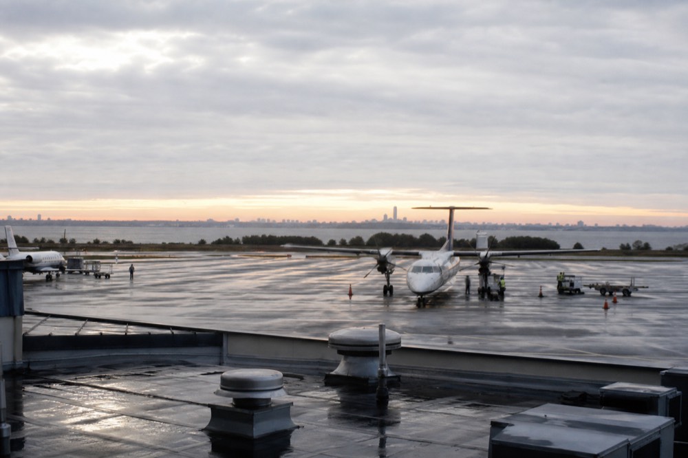Small regional airport apron at sunrise, a turboprop parked on wet concrete, distant ground-crew silhouettes, flat grey sky above a low terminal roof