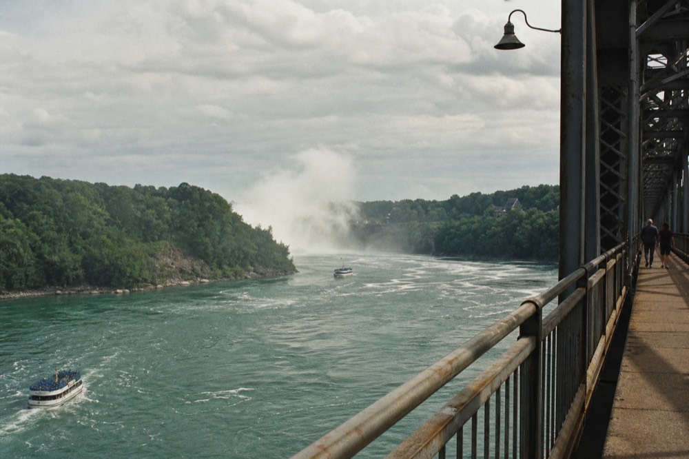 Pedestrian walkway on a steel arch bridge above a fast green river, painted railings in the foreground, a distant mist plume rising between wooded banks under overcast light