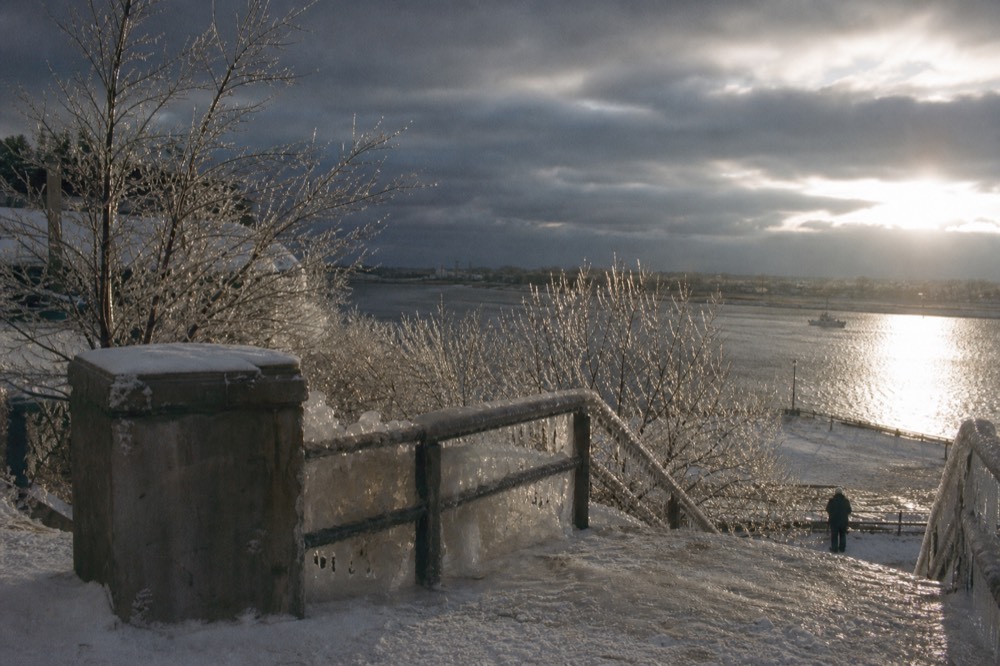 Ice-crusted railings and frozen spray coating bare trees at a river overlook in winter, pale sun low behind cloud, packed snow on worn stone steps