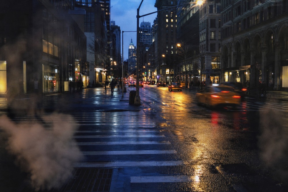 Manhattan avenue at dusk from a sidewalk corner, wet crosswalk stripes underfoot, cab taillights streaking between tall dark buildings and lit shop windows