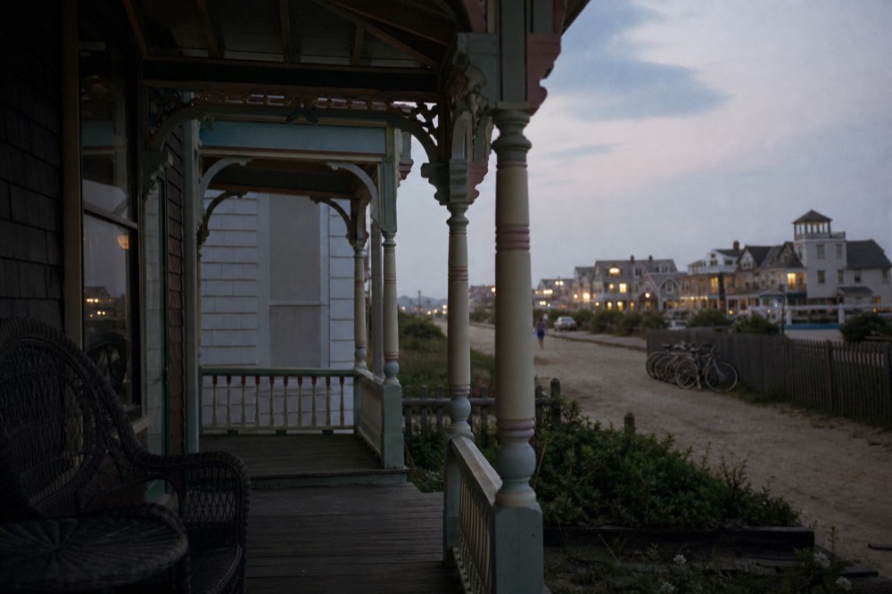 Victorian seaside porch with turned columns and pastel trim, wicker chairs deep in shade, sandy street beyond and bicycles leaning on a fence