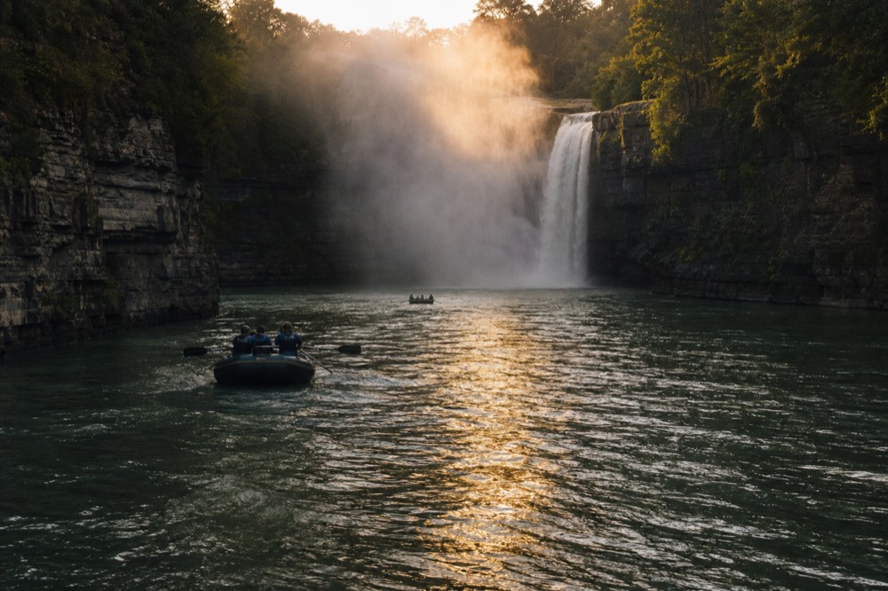 Wide river gorge below a waterfall at dusk, dark wet cliff walls on both sides, a column of mist catching the last light above deep green water